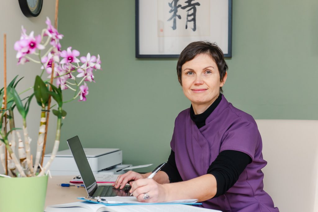 Lucy Clarke in her clinic using a laptop, smiling at the camera while reviewing functional health assessments