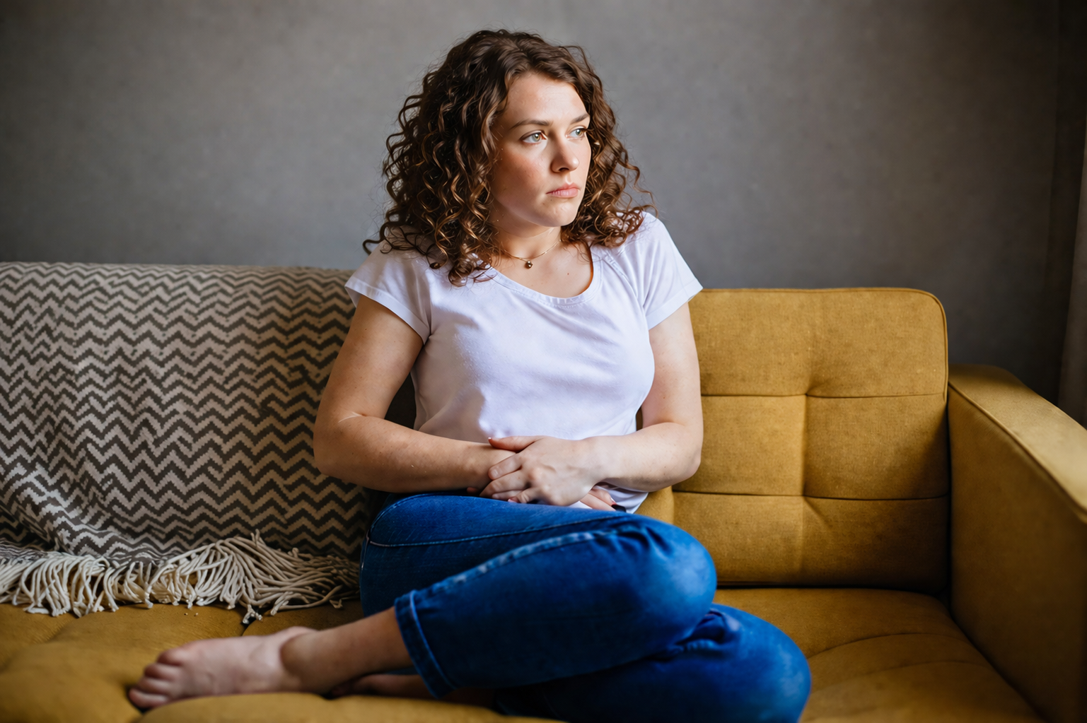 Woman with brown curly hair sitting curled up on a mustard sofa, holding her stomach with a pained expression, illustrating abdominal discomfort, bloating, or digestive pain in a home setting.